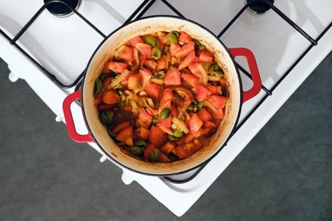 Red cast iron pot sits on white stove. Inside pot are chopped tomatoes, onion, capers and green