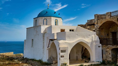 White church with blue dome by the sea, surrounded by old stone walls under a blue sky, Church of