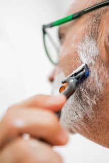 Close-up of a mature man shaving in the bathroom. The man, wearing glasses, uses a razor across a
