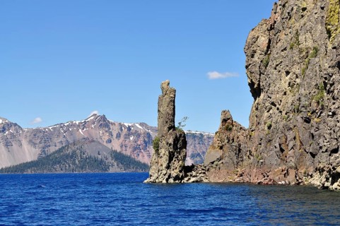 Rock formation with mountain backdrop on a clear lake under bright blue sky, Crater Lake National
