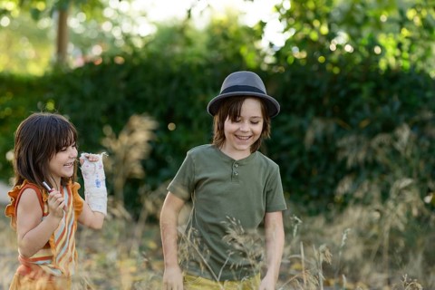 Two children joyfully explore a garden, surrounded by lush greenery. One wears a hat, while the