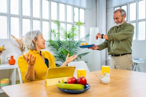 Senior man serving pastries to his wife while using digital tablet sitting on the living room She