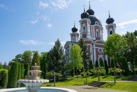 Orthodox church with red and white accents surrounded by a well-kept garden with a fountain, blue