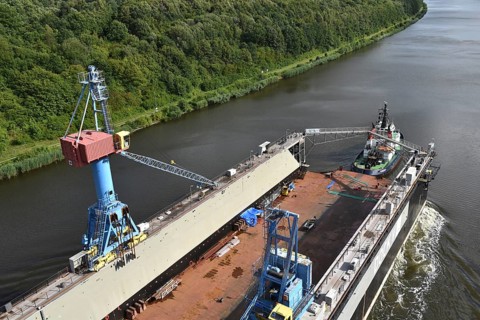 A tug and barge on a river, with forest and transport equipment on deck, tugboat Bremerhaven pulls