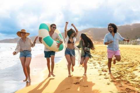 Joyful diverse young friends running barefoot on the sand, laughing and celebrating during a sunny