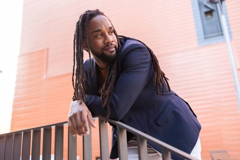 A cuban business man with dreadlocks, wearing a navy blazer, leans thoughtfully on a railing