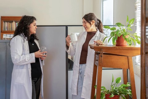 Two young female scientists in white lab coats chatting during a break, one holding a mug and the