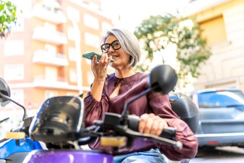 Active senior woman with grey hair sitting on a moped on a city street, speaking into a smartphone,