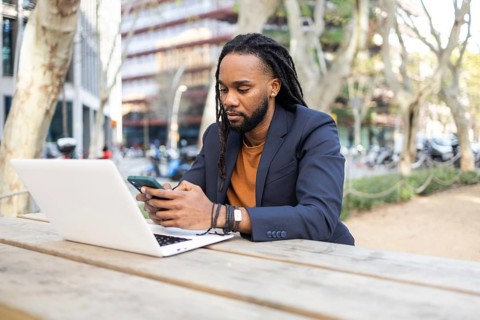A cuban businessman in a blazer uses a smartphone and laptop while sitting on a wooden bench