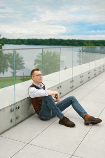 Middle-aged man sits on terrace overlooking lake. He leans back against glass barrier. Calm day