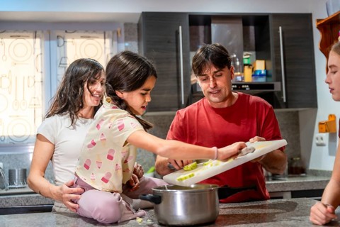Parents and two daughters cooking together, with a young girl sitting on the counter helping father