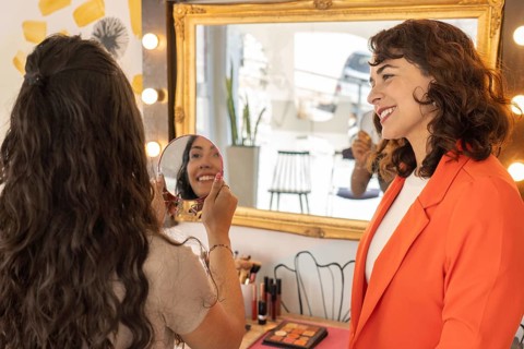 Rear view of a client smiling while examining her completed makeup in a hand mirror at a beauty