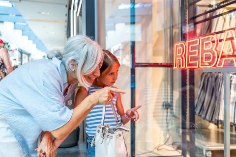 Grandmother and granddaughter are happily window shopping at a mall, looking at a clothing store