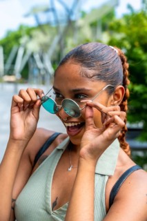 Biracial young woman lowering round sunglasses, looking playfully with a wide smile, expressing