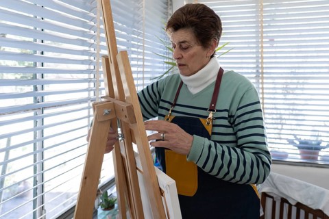 A senior female artist in a denim apron leans over her wooden easel to tighten a screw and secure a