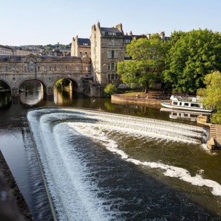 River with small waterfall and bridge, surrounded by old buildings, Bath