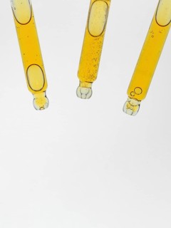 Three glass pipettes for cosmetics filled with amber liquid are suspended against a clear white