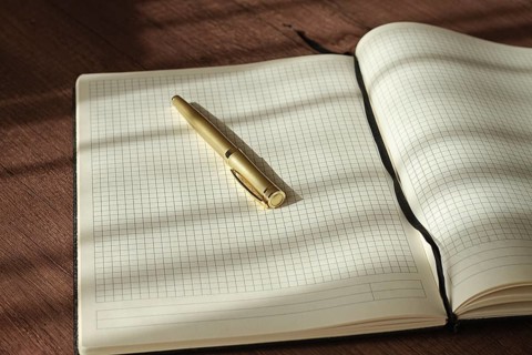 A notebook with blank grid pages is open on a wooden table. A pen rests on the notebook as sunlight