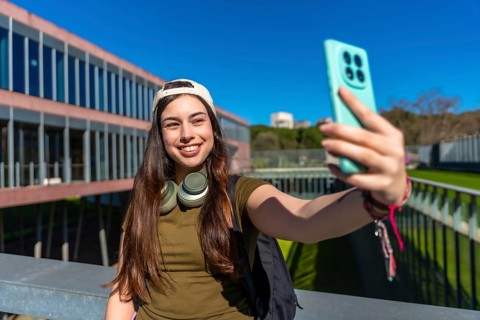 Young female college student with headphones and backpack smiling while holding a smartphone and