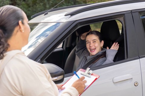Young woman student raising hands and laughing joyfully while driving instructor writes notes on