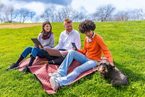 Three students are using electronic devices while relaxing on a blanket in a park with their dog