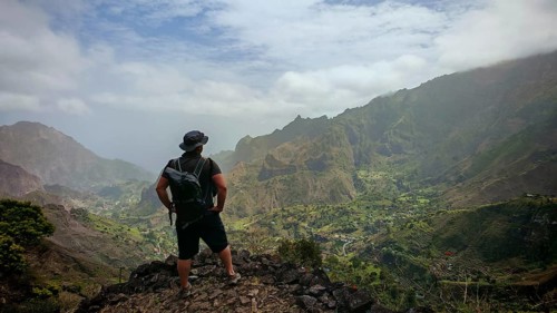 Male person with backpack stands on a rock and looks into the valley surrounded by green mountains