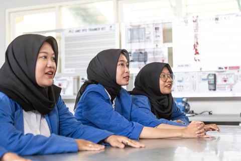 Three Asian Muslim students in hijabs and blue uniforms sit attentively at a classroom desk,