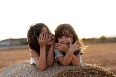 Two children enjoy a playful moment on a hay bale during sunset, smiling and covering their faces