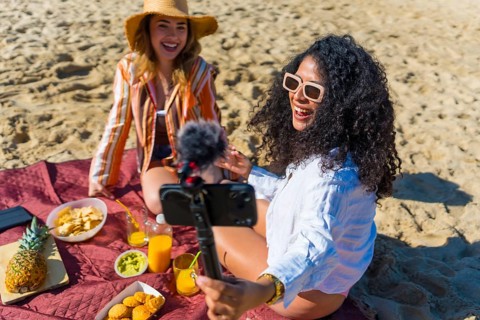 Two cheerful young women, one latina with curly hair holding a smartphone on a tripod with a