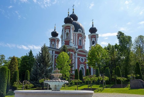 Orthodox church with red and white accents surrounded by trees against a blue sky with a fountain