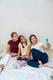 Three happy young sisters sharing a moment, taking a selfie with a smartphone, and relaxing on a
