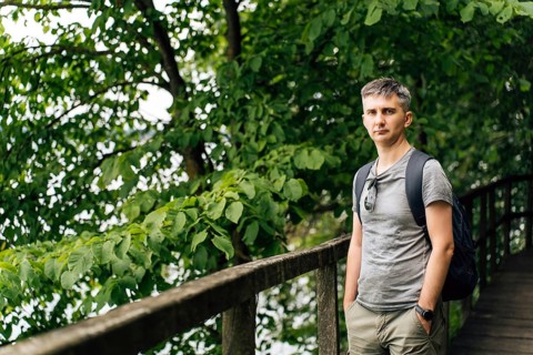 A man walks along a wooden forest walkway surrounded by dense green foliage. He wears a gray