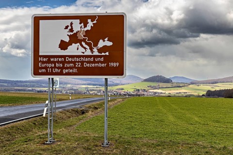 Road sign with map of Europe and text against green landscape and cloudy sky, The Point Alpha