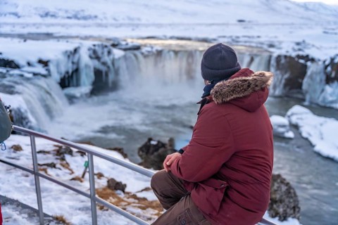 A man hiker sitting on the frozen Godafoss waterfall at sunset in winter, Iceland