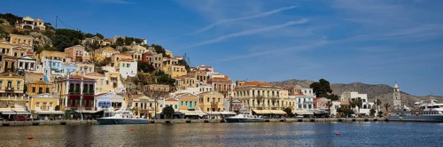 Colourful houses and boats line a tranquil coastline under a clear sky, Symi Town, Symi,