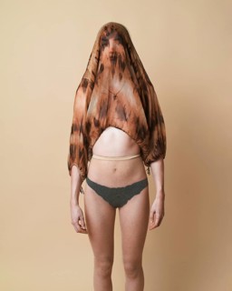 A young woman stands against a beige background in a studio, wearing a sheer top over her head