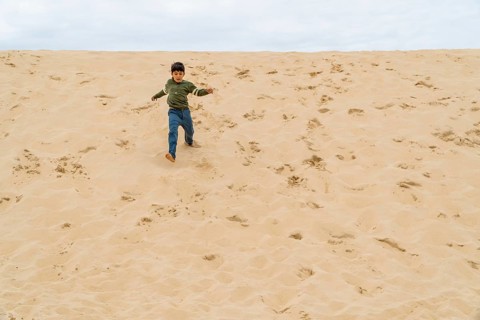 A young boy in casual clothing joyfully runs down the dune of Pilat under an overcast sky in France