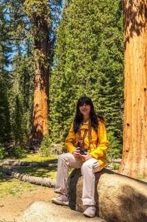 Woman wearing a yellow jacket and holding a camera while sitting on a rock, smiling at the camera