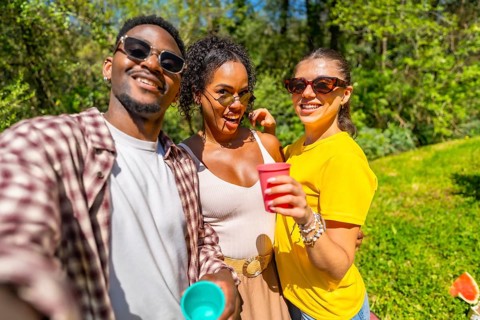 Three cheerful friends enjoying a picnic, taking a selfie and holding colorful cups on a sunny day