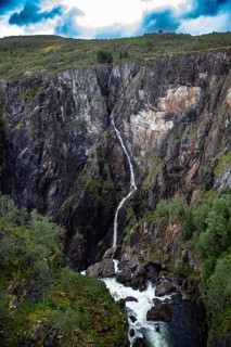A high waterfall flows through rugged cliffs surrounded by thick vegetation, under a cloudy sky,
