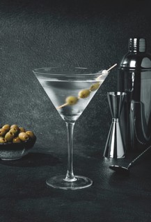 A drink is prepared in a bar setting. A martini glass is filled with clear liquid and has olives on