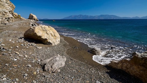 A quiet beach with pebbles and rocks, surrounded by crystal clear water and mountains in the