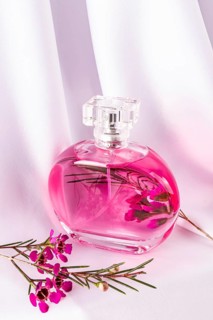 A sleek, pink-hued perfume bottle rests on elegant white fabric, surrounded by vibrant pink flowers