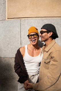 A gay couple joyfully embraces while holding hands outdoors. They wear stylish casual outfits and