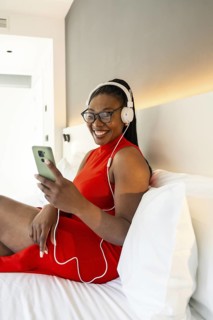Smiling businesswoman wearing headphones using smartphone on hotel bed, enjoying music during