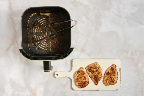 Air fryer sits on counter with three cooked pork chops on white cutting board nearby. Kitchen