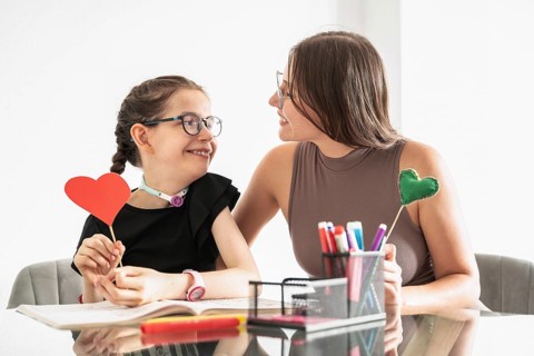 Disabled girl with a tracheostomy, sitting with sister who supports her. They are engaged in a