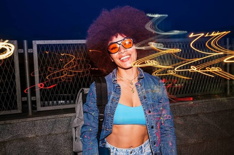 A cheerful African descendent woman with afro hair, wearing orange sunglasses and a denim jacket,