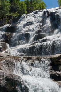 A majestic waterfall flows in several cascades over rocks, surrounded by trees in sunny weather,