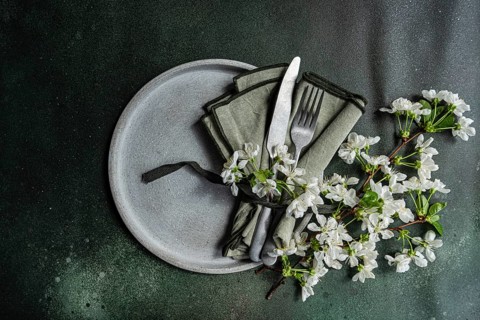 A minimalist clay plate with green napkin, silverware, and white blossoms on a dark textured
